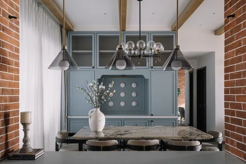 A Mumbai apartment dining space with a timber table, dark chairs, blue fitted cabinets, brick accents and pendant lights